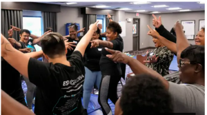 A group of students training in a new self-defense for Women of Color workshop. The women are seen from the side and rear, with their hands raised in a confident, protective gesture.