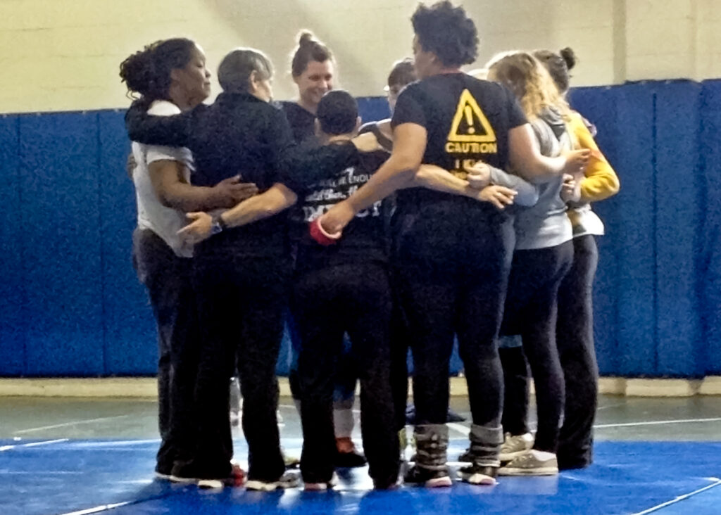 Women taking an empowerment self-defense class huddle together for support and encouragement before a round of practice scenarios. The women are young and old, White and and Black, coming together as sisters to empower one another and end violence.
