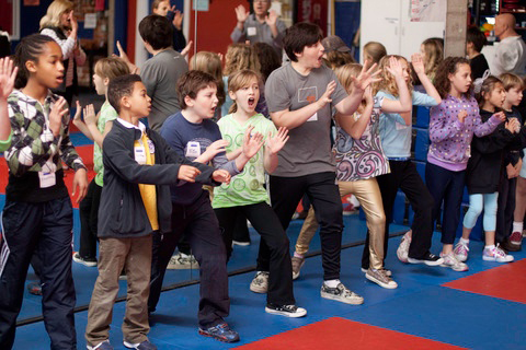 Photo in landscape orientation of a group of children learning empowerment self-defense. Their hands are raised in the stop sign gesture and they are practicing their loud-voiced "Stop!" commands.